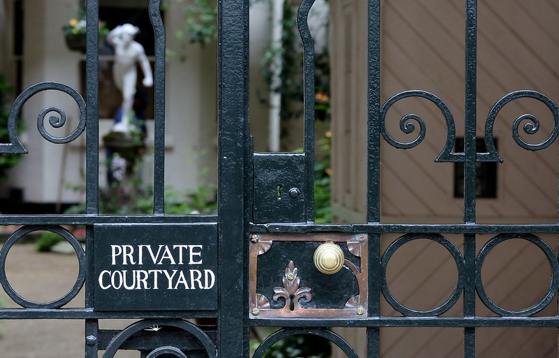 Image shows a locked gate with a sign reading "Private Courtyard." In the background is a blurry but probably lovely scene with foliage and an elegant statue in a Classical style.